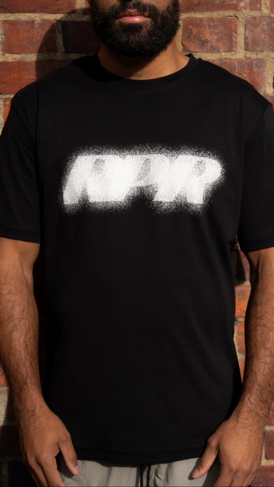 Model wearing black t-shirt with white spray logo graphic, standing against brick wall, paired with grey track pants